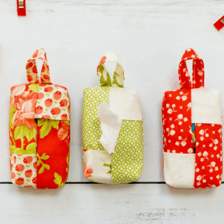 Three handmade fabric pouches with handles, each in patchwork red, green, and white patterns, are displayed on a white surface alongside sewing clips, fabric scraps, spools of thread, and inspiring travel tissue holder designs to sew.
