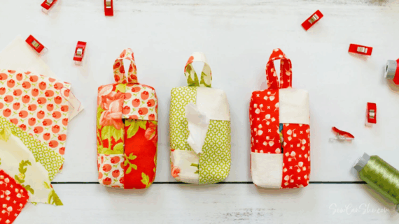 Three handmade fabric pouches with handles, each in patchwork red, green, and white patterns, are displayed on a white surface alongside sewing clips, fabric scraps, spools of thread, and inspiring travel tissue holder designs to sew.