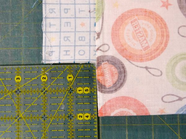 Two pieces of patterned fabric are aligned on a green cutting mat with yellow grid lines. A clear, yellow-marked quilting ruler is placed along the fabric edge&mdash;perfect for learning how to sew box corners precisely.