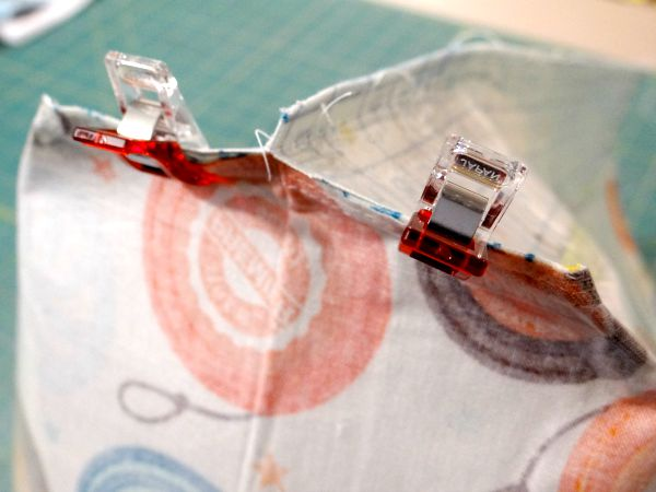 Two fabric pieces with colorful patterns are clipped together at the edge using two transparent red sewing clips, preparing for box corner sewing. A green cutting mat is visible in the background.