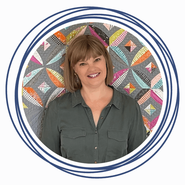 Smiling woman with straight, shoulder-length light brown hair, wearing a dark green button-up shirt, stands in front of a colorful geometric quilt and blue concentric circles&mdash;ready to move the needle at the upcoming quilting summit.
