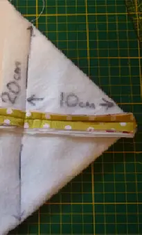 A piece of white fabric folded into a triangle with &ldquo;20 cm&rdquo; and &ldquo;10 cm&rdquo; marked in black, beside a green polka-dotted strip, on a green and yellow grid cutting mat&mdash;perfect for sewing pouches or learning how to Sew Box Corners.