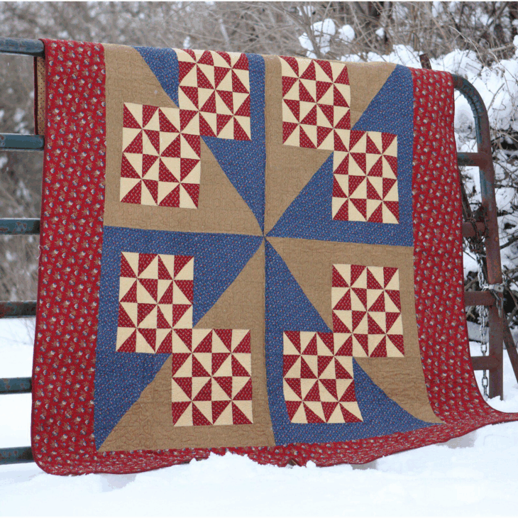 250th Anniversary Pattern Bundle - Limited Time 8 A colorful quilt with red, blue, and tan geometric pinwheel patterns from a Pattern Bundle hangs over a metal fence outdoors, with snow on the ground and trees in the background.