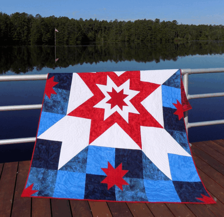 250th Anniversary Pattern Bundle - Limited Time 10 A colorful quilt with an Anniversary Pattern of red, white, and blue stars hangs over a metal railing on a dock, with a calm lake and forested shoreline in the background.