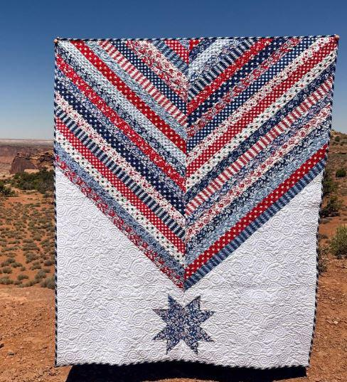 250th Anniversary Pattern Bundle - Limited Time 15 A quilt with red, white, and blue chevron stripes and patterns, plus a star design at the bottom, is held outdoors in a desert landscape under a clear blue sky—perfect for celebrating with an Anniversary Pattern Bundle.