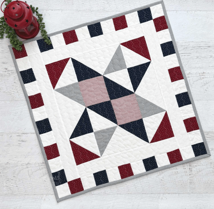 250th Anniversary Pattern Bundle - Limited Time 17 A square quilt from the Anniversary Pattern Bundle with a geometric pattern in red, white, and dark blue, featuring triangles and rectangles. The border alternates between red and blue squares, with a red lantern and greenery in the top left corner.
