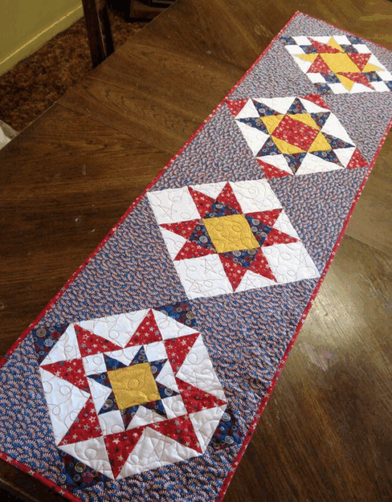 250th Anniversary Pattern Bundle - Limited Time 20 A patriotic table runner on a wooden table, featuring quilted star patterns in red, white, blue, and yellow fabrics—an ideal Anniversary Pattern—bordered with red edging and set against a blue and white patterned background.