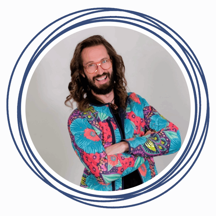 A smiling person with long curly hair and glasses wears a colorful floral-patterned jacket. They stand with arms crossed inside a circular blue-and-white border&mdash;a perfect fit for the inspiring Move the Needle quilting summit.