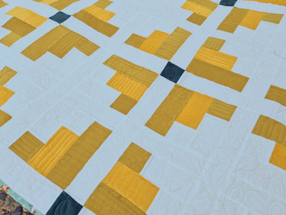 A close-up view of a Mustard Seed Quilt featuring a geometric pattern with yellow and gold rectangles on a white background, intersected by small dark squares and decorative white stitching&mdash;perfect for your next Monthly Make & Take session.