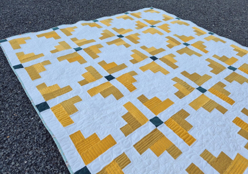 A handmade quilt laid on gravel, featuring a geometric pattern of yellow and white shapes with small dark green squares at some intersections. The quilt has a grid-like, symmetrical design.