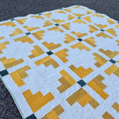 A handmade quilt laid on gravel, featuring a geometric pattern of yellow and white shapes with small dark green squares at some intersections. The quilt has a grid-like, symmetrical design.