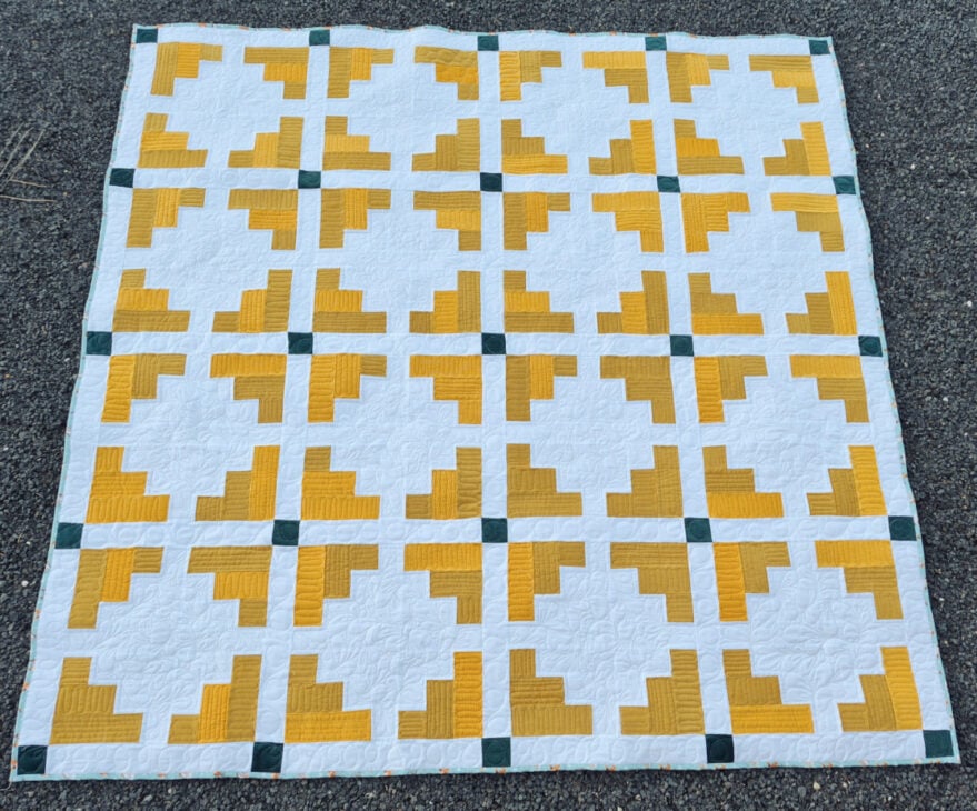 A handmade quilt with a geometric pattern of yellow and white shapes forming squares, separated by small green squares at the intersections, laid out on a textured dark surface.