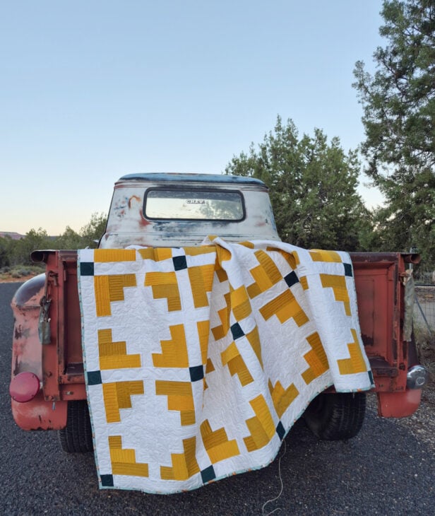 A yellow and white patchwork quilt is draped over the tailgate of an old, rusty red pickup truck parked on a rural road, with trees and a clear sky in the background.