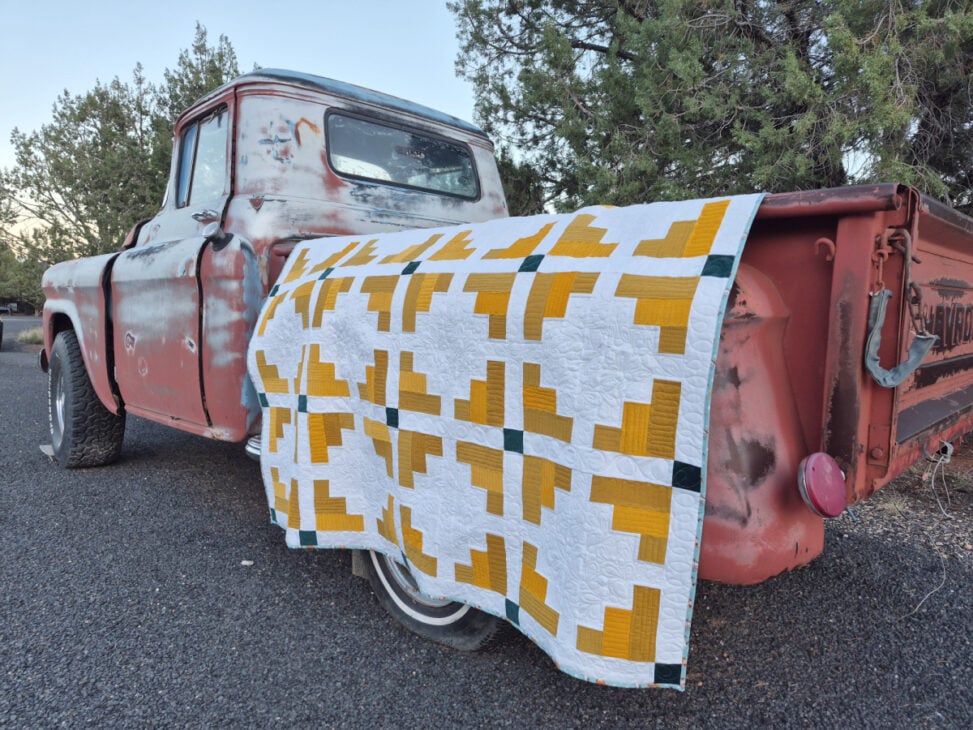 A weathered red pickup truck is parked outdoors with a Mustard Seed Quilt, known from our Monthly Make & Take, draped over its tailgate. Trees and asphalt are visible in the background.