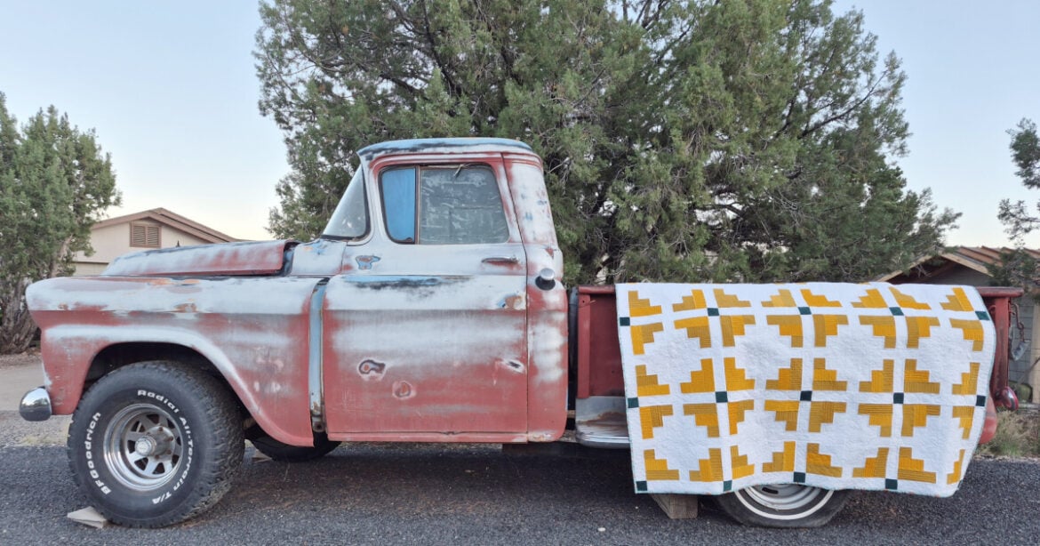 An old, weathered red pickup truck is parked by trees and houses. A white, yellow, and grey geometric-patterned Mustard Seed Quilt, featured in our Monthly Make & Take, hangs over the truck bed&rsquo;s side.