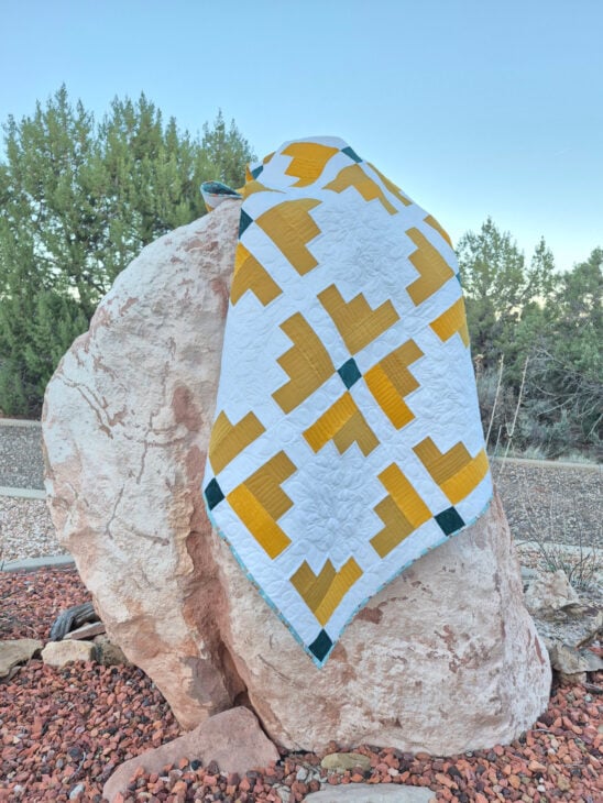 A yellow, green, and white Mustard Seed Quilt with a geometric pattern drapes over a large, light-colored rock outdoors, surrounded by small red rocks and trees in the background.