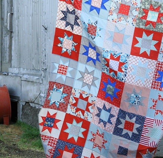 A colorful patchwork quilt with red, white, and blue star patterns reminiscent of the American Flag hangs outdoors near a weathered wooden building and a red barrel.