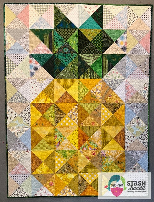 A quilt made of triangle patches arranged in a heart shape, with green fabrics forming the heart at the top and yellow below, creating a gradient effect. Inspired by the pineapple quilt block, it features a variety of patterns and textures.
