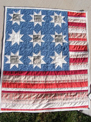 A handmade quilt inspired by the American Flag, featuring blue and white star patterns in the upper left and red, white, and pink stripes on the right and bottom, laid out on a concrete surface.