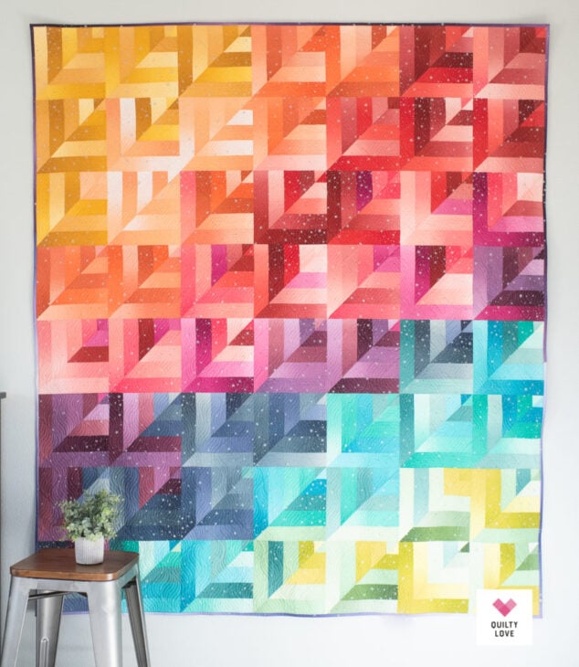 Rainbow Quilt Patterns That Turn Scraps Into Something Stunning 3 A vibrant rainbow quilt hangs on a wall, featuring diagonal geometric patterns in a gradient of warm reds, oranges, and yellows that blend into cool greens, blues, and purples on the bottom right. A plant sits on a stool nearby.