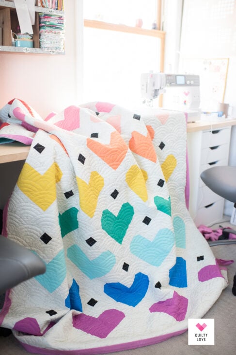 Rainbow Quilt Patterns That Turn Scraps Into Something Stunning 1 A colorful rainbow quilt with heart patterns draped over a chair in a bright sewing room, with a sewing machine and fabric visible on the desk in the background.