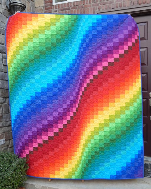 Rainbow Quilt Patterns That Turn Scraps Into Something Stunning 6 A colorful rainbow quilt hangs outside, featuring a vibrant wave pattern made of small squares in a gradient from red to purple, with the colors blending smoothly across the fabric.