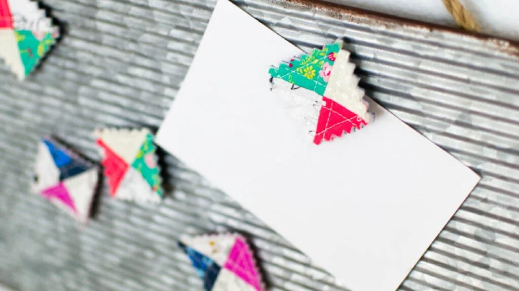 A blank white card rests on a ribbed metal surface, held by a colorful, triangular magnet covered in crumb quilt patchwork fabric. Other similar magnets are visible nearby.