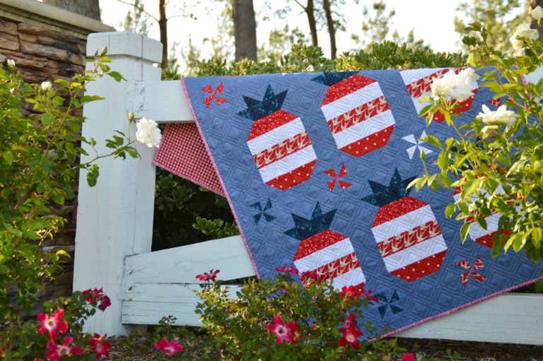 A quilt with red, white, and blue pineapple patterns, reminiscent of the American Flag, hangs over a white wooden fence surrounded by blooming flowers and greenery on a sunny day.