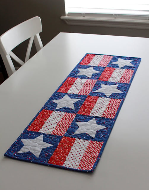 A rectangular table runner with a patchwork design inspired by the American Flag, featuring red, white, and blue fabrics decorated with white stars and various patterns, is placed on a white table next to a white chair.