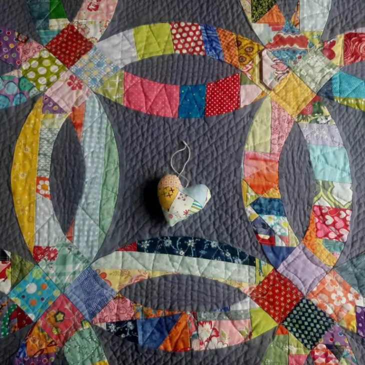 A colorful patchwork double wedding ring crumb quilt with overlapping rings and a small fabric heart ornament placed on top. The quilt features various patterned fabrics in bright, vibrant colors.