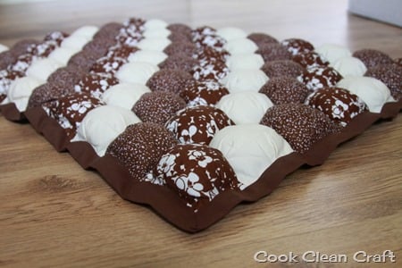 A handmade, padded bubble quilt crafted with Quilt Sewing techniques features alternating rows of brown, patterned brown, floral brown, and white squares. Displayed on a wooden floor, "Cook Clean Craft" is inscribed in the bottom right corner.