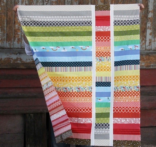 A person holds up a colorful quilt made of horizontal fabric strips in various patterns and colors, including reds, blues, greens, and yellows, with two vertical white strips near the center&mdash;perfect inspiration for Quilt Patterns enthusiasts.