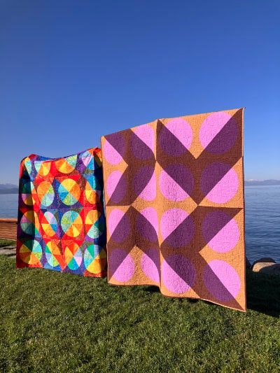 Two colorful quilts, crafted with scrap quilting techniques, are displayed outdoors on green grass near a body of water. One showcases bright geometric patterns; the other features large overlapping pink and brown shapes beneath a clear blue sky.