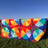 A colorful scrap quilt with curved, circular geometric patterns in bright hues is draped over a wooden bench on green grass, with a blue sky and water in the background.
