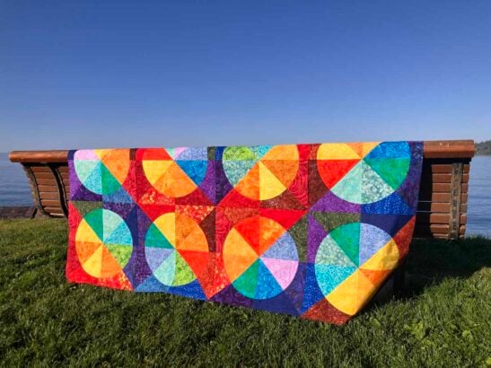 A colorful scrap quilt with curved, circular geometric patterns in bright hues is draped over a wooden bench on green grass, with a blue sky and water in the background.