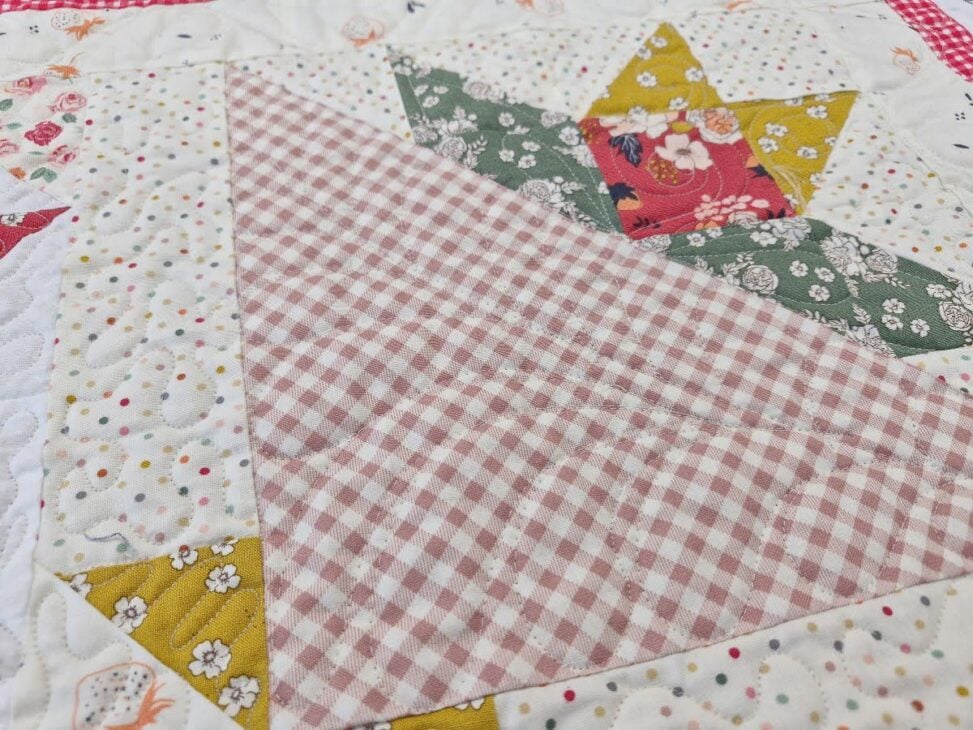 June 2026 BOM 2 A close-up view of a quilt block from the June 2026 BOM, featuring a pink and white gingham triangle, surrounded by floral and polka dot fabrics in white, yellow, and green. The quilt has a patchwork and stitched texture.