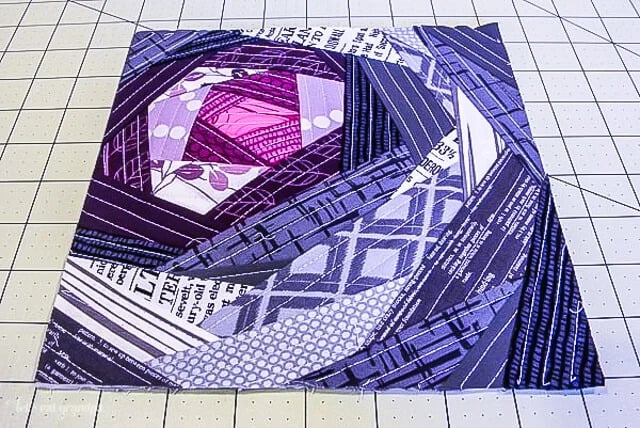 A quilt block featuring various purple and violet fabric patterns, arranged in a geometric spiral design, lies on a gridded cutting mat&mdash;perfect inspiration for Quilt Patterns or Quilt As You Go techniques.