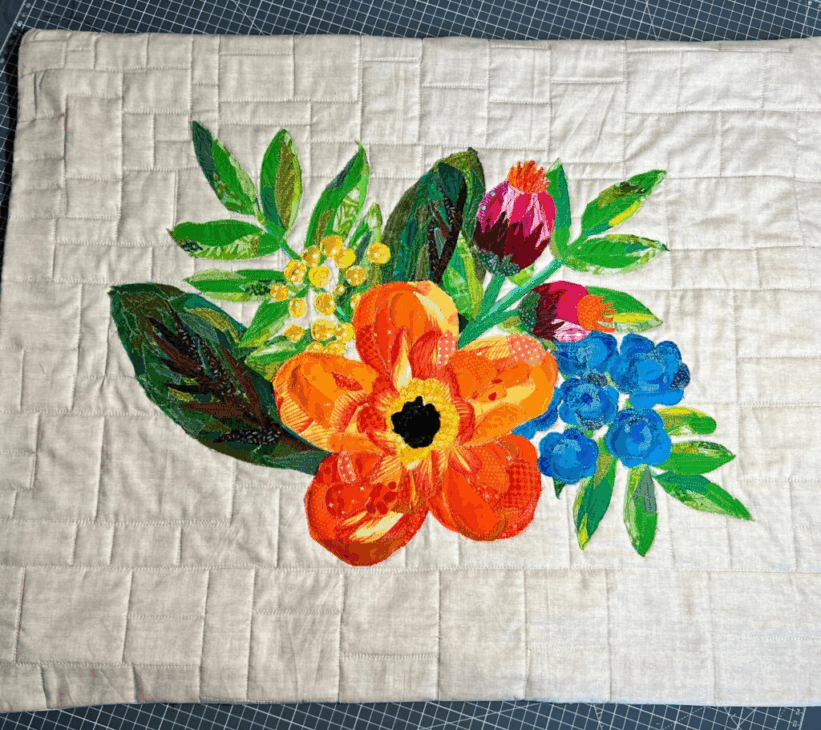 A fabric collage appliqu&eacute; of a vibrant floral bouquet, featuring an orange poppy, blue berries, and green leaves, on a quilted, light-colored background&mdash;perfect inspiration for unique quilt patterns.