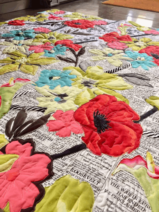 A colorful fabric collage quilt with large, vibrant floral patterns is laid out on the floor. The background features black-and-white newspaper print, giving inspiration for unique fabric collage quilt patterns.