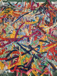 A colorful abstract textile artwork, inspired by quilt patterns, features overlapping strips and threads in vibrant hues&mdash;red, yellow, blue, green, and purple&mdash;creating a visually dynamic and textured fabric collage composition.