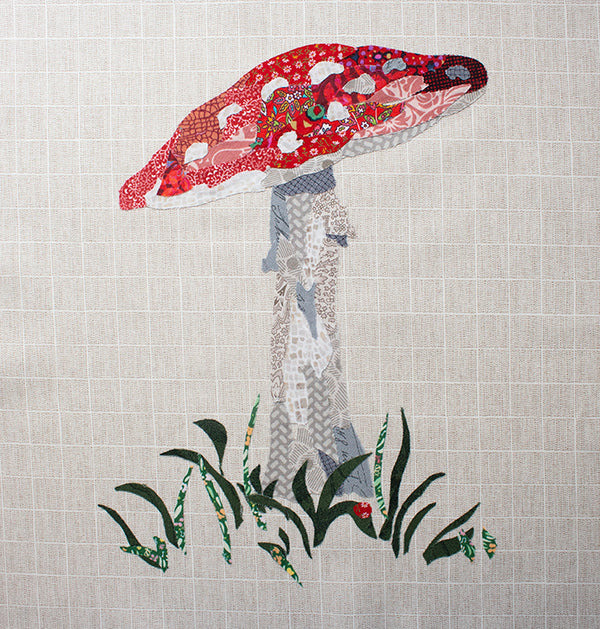 A fabric collage art piece depicts a red and white mushroom with a tall gray stem, surrounded by green grass on a beige, grid-patterned background&mdash;perfect inspiration for quilt patterns or free ideas.