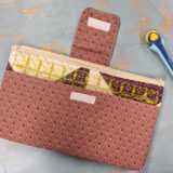 A patterned fabric pouch crafted from a Notion Bag Pattern with a flap and zipper holds a clear quilting ruler on a gray surface next to a blue and yellow rotary cutter.