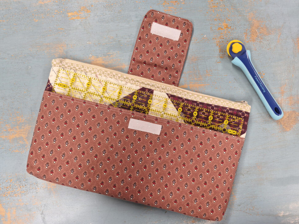 A patterned fabric pouch crafted from a Notion Bag Pattern with a flap and zipper holds a clear quilting ruler on a gray surface next to a blue and yellow rotary cutter.