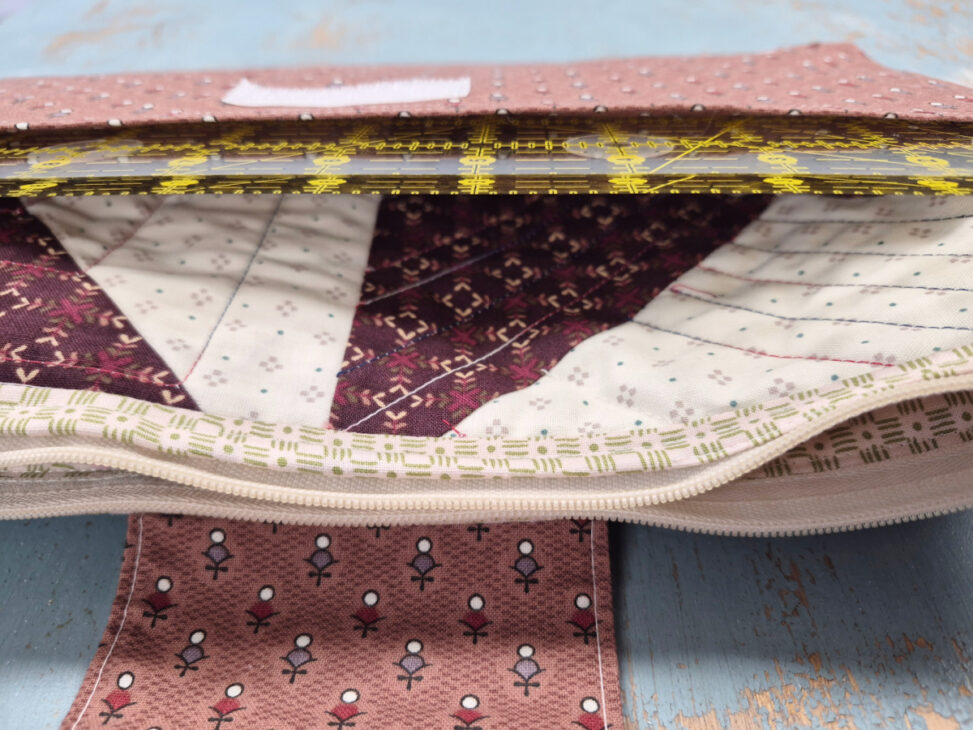 A quilted fabric pouch made with a Notion bag pattern, partially unzipped to reveal a ruler inside. The pouch features pink, white, and purple geometric and floral patterns on a blue wooden surface—perfect for your next sewing day.