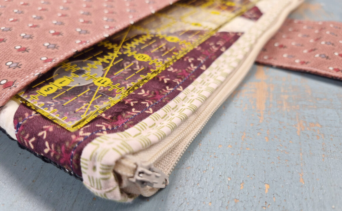 A close-up of a fabric Notion Bag with a zipper, partially open to reveal a clear yellow quilting ruler and purple patterned fabric inside. The pouch has a pink exterior with small white patterns—perfect for organizing your sewing essentials.