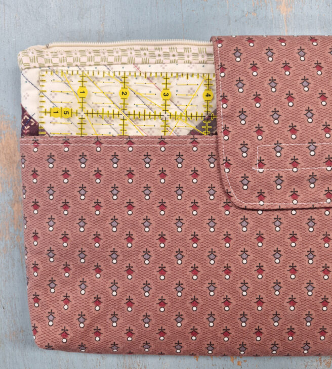 A pink patterned Notion Bag with a snap button lies on a blue textured surface. A quilting ruler and sewing fabric pieces are partially visible, sticking out from the pouch's open pocket.