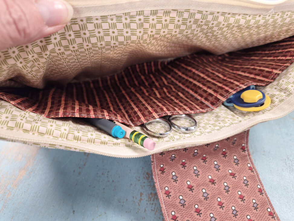 A close-up of an open notion bag reveals pockets holding a pencil, a pen, a pair of scissors, and a button, with patterned fabric lining and a snap closure visible—perfect for keeping your sewing essentials organized.