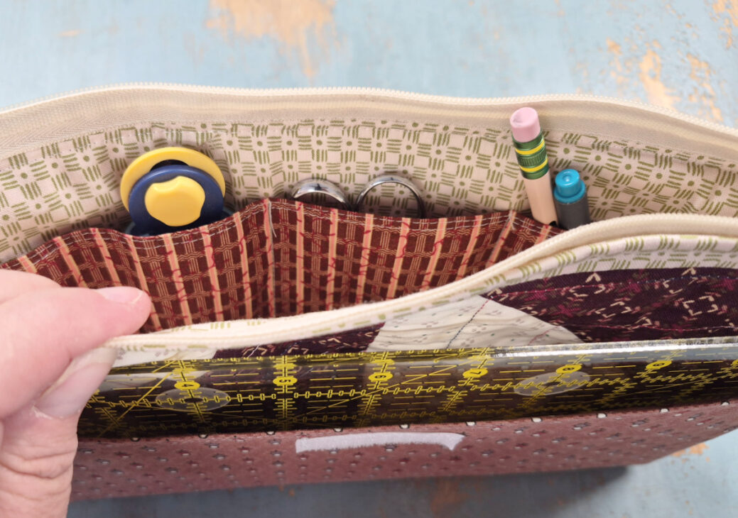 A hand holds open a zippered notion bag with various compartments, revealing contents including a rotary cutter, scissors, a pencil, a pen, a ruler, and other crafting supplies—perfect for an organized sewing day.