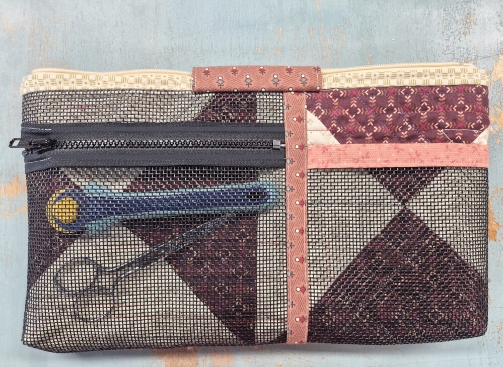 A quilted fabric pouch with a black mesh front, zipper pocket, and patchwork design holds a blue rotary cutter and small scissors, visible through the mesh—ideal for storing tools on Sewing Day or using as a Notion Bag Pattern.