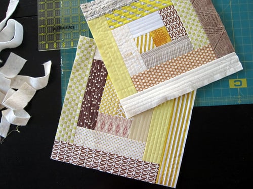 Two quilt blocks made from strips of fabric in yellow, brown, white, and beige patterns are laid on a cutting mat next to fabric scraps and a clear ruler&mdash;perfect inspiration for Quilt As You Go or exploring new Quilt Patterns.